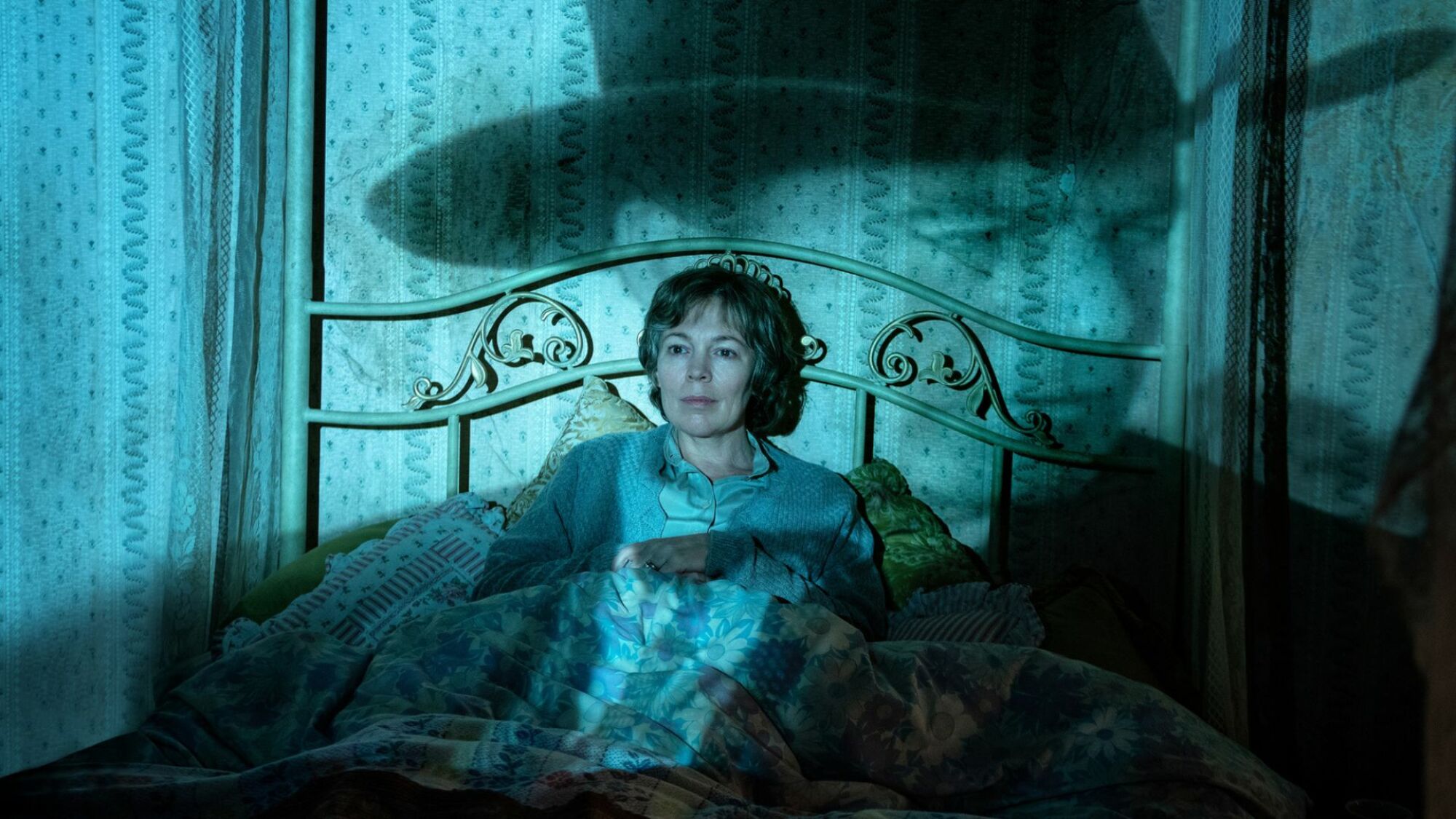 A middle-aged woman sits on a bed, a projection of a cowboy plays over her.